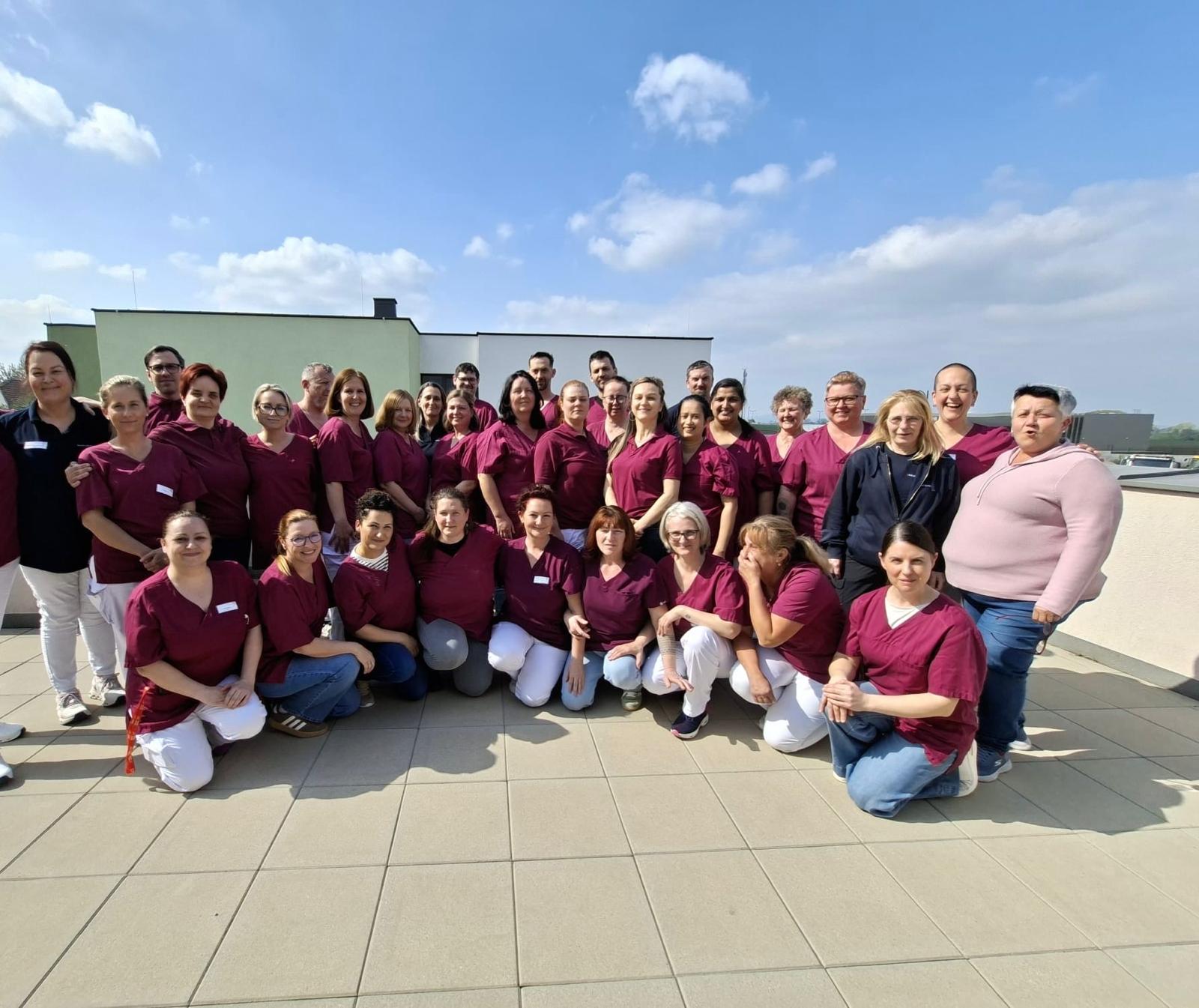 Ein großes Team von etwa 40 Personen in roten Oberteilen steht auf einem Dach mit Wolken am Himmel. Einige Personen lächeln, während andere in die Kamera schauen. Die Gruppe scheint in einem professionellen Umfeld, möglicherweise im Gesundheitswesen, zu sein.