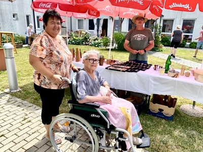 Eine ältere Frau im Rollstuhl steht neben einer lächelnden Frau an einem Marktstand.