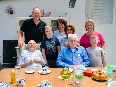 Eine Gruppe von sechs Personen steht und sitzt um einen Tisch im Freien. Auf dem Tisch stehen Getränke und Gebäck. Zwei ältere Menschen sitzen im Vordergrund, während die anderen Personen lächeln und in die Kamera schauen. Die Umgebung ist hell und freundlich.