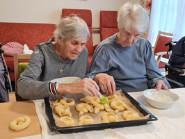 Zwei ältere Frauen sitzen an einem Tisch und backen. Eine Frau streicht einen Teigling mit einem Pinsel ein, während die andere neben ihr mit einem Schüssel Mehl sitzt. Auf einem Blech liegen mehrere geformte Teigstücke. Im Hintergrund sind rote Stühle sichtbar.