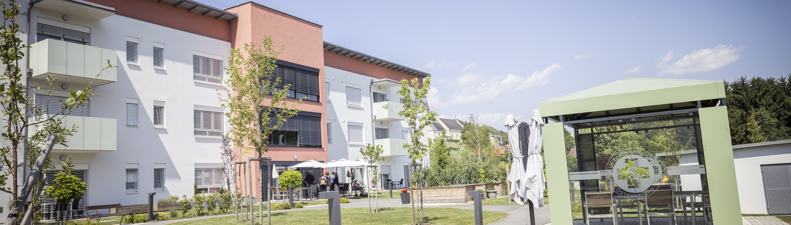 Ein modernes Gebäude mit bunten Fassaden und Balkonen. Im Vordergrund eine gepflegte Grünfläche mit Wegen und Bäumen. Auf der rechten Seite steht ein Pavillon mit Sitzgelegenheiten. Der Himmel ist klar und sonnig, was eine einladende Atmosphäre schafft.