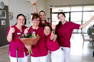 Fünf fröhliche Mitarbeiterinnen in roten Poloshirts präsentieren einen Blumenstrauß.