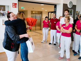 Zwei Frauen umarmen sich, eine hält einen Blumenstrauß. Das Team schaut zu.
