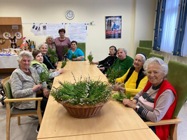 Eine Gruppe älterer Frauen sitzt an einem Tisch mit grünen Zweigen in der Hand.