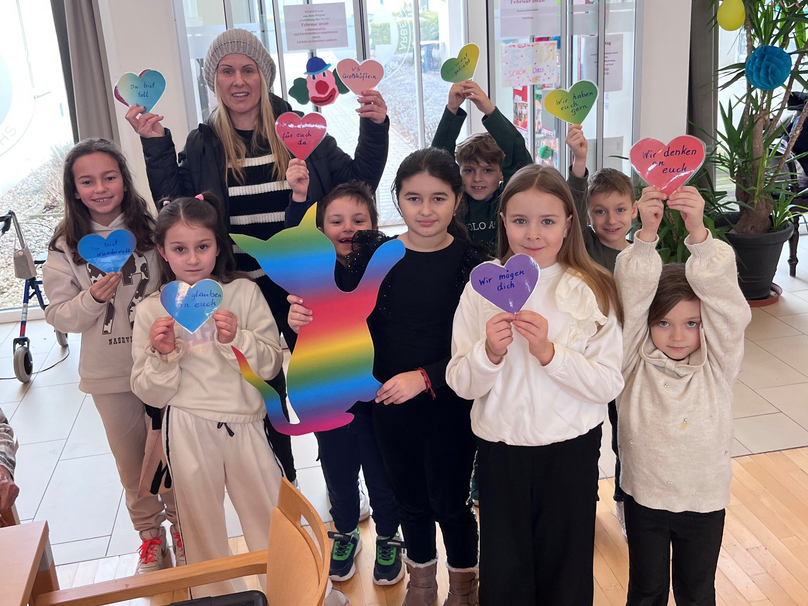 Gruppe von Kindern zeigt bunte Herzchen und eine Regenbogenfigur in einem Raum.