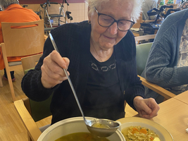 Eine ältere Frau schöpft Suppe aus einem großen Topf in einen Teller.