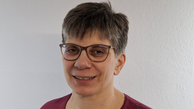 Frau mit Brille lächelt, trägt ein rotes Oberteil und steht vor einer weißen Wand.