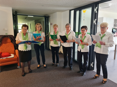 Sechs Frauen singen gemeinsam, tragen weiße Blusen und grüne Schals.