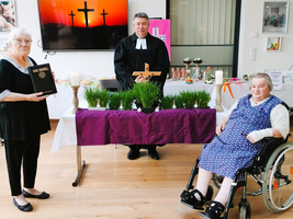Eine Frau hält die Bibel, ein Pfarrer steht mit einem Kreuz, eine weitere Frau im Rollstuhl.
