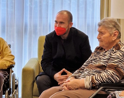Ein Mann mit roter Maske spricht mit zwei älteren Frauen in einem Aufenthaltsraum.