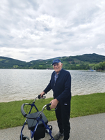 Ein älterer Mann steht neben einem Rollator am Ufer eines Sees.