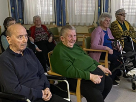 Ältere Menschen sitzen in einem Raum und lächeln freundlich.