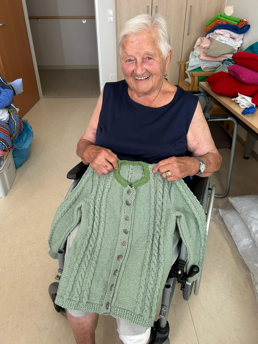 Eine lächelnde Frau im Rollstuhl hält einen grünen handgestrickten Pullover hoch.