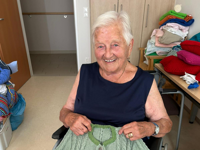 Eine lächelnde Frau im Rollstuhl hält einen grünen handgestrickten Pullover hoch.