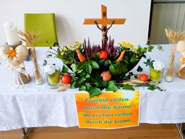 Tischdekoration mit Früchten, Blumen und einem religiösen Kreuz. Schriftzug: „Durch Liebe reifen Menschen.“