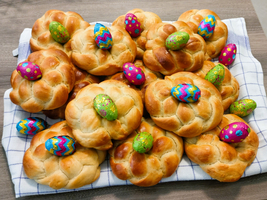 Eine Anordnung von frisch gebackenen, geflochtenen Brötchen auf einem karierten Küchenhandtuch, dekoriert mit bunten Schokoladeneiern in lebhaften Farben. Die Brötchen sind goldbraun und glänzend, während die Eier in verschiedenen Mustern und Farben leuchten.