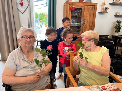 Seniorinnen halten Rosen, umgeben von lächelnden Kindern in einem Innenraum.