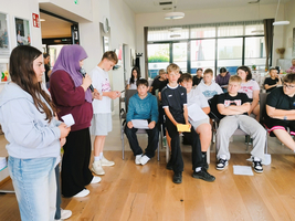 Gruppendiskussion in einem Raum mit vielen Schülern. Eine Schülerin mit einem lila Hijab spricht ins Mikrofon, während andere Schüler in Stühlen sitzen und zuhören. Der Raum hat große Fenster und ist hell beleuchtet.