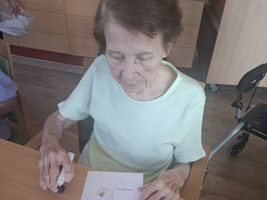 Eine ältere Frau schreibt auf ein Blatt Papier in einem sonnigen Raum.