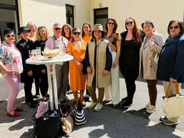 Eine Gruppe von 14 Personen steht um einen Stehtisch im Freien. Sie lächeln und posieren für ein Foto. Einige tragen Sonnenbrillen und verschiedene Outfits, die von leger bis elegant reichen. Im Hintergrund sind Wände eines Gebäudes sichtbar.