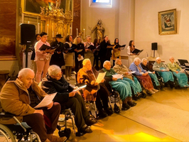 Eine Gruppe von Senioren sitzt in einer Halle, einige im Rollstuhl. Sie halten Liederblätter in den Händen. Im Hintergrund steht ein gemischter Chor aus mehreren Personen, die in verschiedenen Kleidern singen. Der Raum ist hell erleuchtet und hat religiöse Kunstwerke.