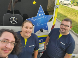 Drei Personen stehen vor einem Fahrzeug mit der Aufschrift 