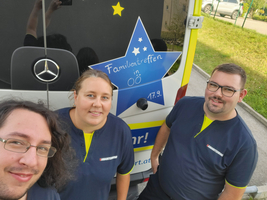 Drei Personen stehen vor einem Fahrzeug mit der Aufschrift 