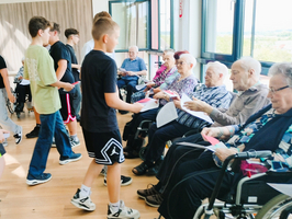Eine Gruppe von Jugendlichen verteilt Farbkarten an ältere Menschen, die in Rollstühlen sitzen. Sie befinden sich in einem hellen Raum mit großen Fenstern, die einen Blick auf die Landschaft bieten. Die ältere Generation lächelt und interagiert mit den jungen Besuchern.