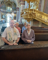 Vier Personen in einer Kirche, sitzen in einer Bank, mit einem Priester vorne.