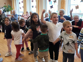 Kinder tanzen fröhlich in einem Raum mit älteren Menschen im Hintergrund.