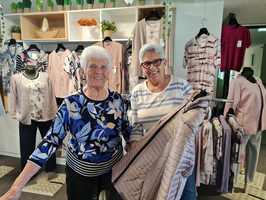 Zwei lächelnde Frauen stehen in einem Geschäft für Bekleidung. Die eine Frau hält eine graue Jacke in der Hand, während die andere in einem bunten Oberteil mit Blumenmuster gekleidet ist. Im Hintergrund sind verschiedene Bekleidungsstücke auf Kleiderständern zu sehen.