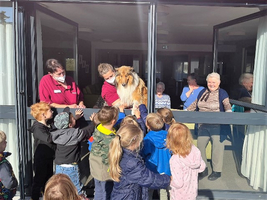 Kinder stehen vor einem Fenster und streicheln einen Hund mit Senioren im Hintergrund.