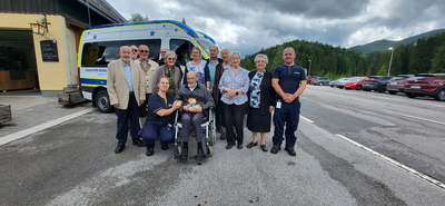 Eine Gruppe von zwölf Personen steht in einem Parkplatz vor einem Krankenwagen. Ein älterer Mann sitzt im Rollstuhl. Die Mitglieder der Gruppe sind unterschiedlich gekleidet, und die Umgebung zeigt eine bewaldete Landschaft unter einem bewölkten Himmel.