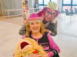 Zwei Frauen tragen bunte Hüte und Brillen, umarmen ein Plüschspielzeug.