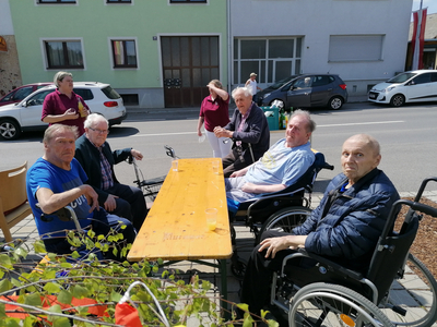 Ältere Menschen sitzen an einem Tisch im Freien, begleitet von Betreuern.