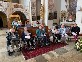 Eine Gruppe älterer Menschen, die in Rollstühlen sitzen, versammelt sich in einer Kirche. Im Hintergrund sind religiöse Kunstwerke und Statuen zu sehen. Einige Personen halten Programme in den Händen, während andere freundlich in die Kamera schauen. Die Atmosphäre wirkt freundlich und gemeinschaftlich.