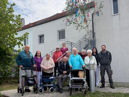 Eine Gruppe von neun Senioren steht vor einem Gebäude. Einige benutzen Rollatoren und andere sitzen in Rollstühlen. Im Hintergrund sind Treppen und ein Baum mit bunten Bändern zu sehen. Es ist Tag und der Himmel erscheint bewölkt.