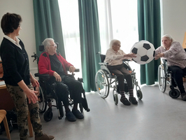 Eine Betreuerin steht neben drei älteren Frauen in Rollstühlen. Eine der Frauen reicht einen großen Fußball weiter, während die anderen lachen und zusehen. Die Szene vermittelt eine freundliche und spielerische Atmosphäre in einer Gemeinschaftseinrichtung.