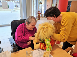 Eine ältere Frau lächelt und interagiert mit einer Puppenspielerin und einer Puppe.