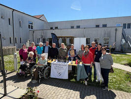Eine Gruppe von Menschen steht im Freien vor einem Gebäude, darunter auch Personen im Rollstuhl. Auf einem Tisch sind Flaschen und Produkte ausgestellt. Es ist sonnig, und die Atmosphäre wirkt herzlich und einladend.