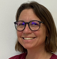 Eine lächelnde Frau mit kurzen, braunen Haaren und einer roten Kleidung steht vor einem weißen Hintergrund. Sie trägt eine Brille mit dunkelblauen Rahmen.