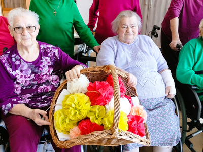 Drei ältere Frauen halten einen Korb mit bunten Papierblumen in einem Gemeinschaftsraum.