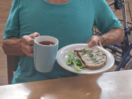 Eine ältere Frau hält eine Tasse und einen Teller mit belegtem Brot.
