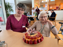 Eine sitzende, ältere Frau mit Brille und grau-weißen Haaren lächelt neben einer Pflegerin in roter Uniform. Vor ihnen steht ein festlicher Obstkuchen, geschmückt mit einer „94“-Kerze. Im Hintergrund sind weitere Senioren und eine hell erleuchtete Umgebung zu sehen.