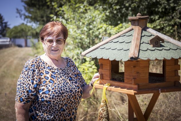 Eine ältere Frau steht neben einem Vogelfutterhaus im Freien.
