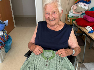 Eine ältere Frau sitzt im Rollstuhl und hält einen grünen Strickpullover.