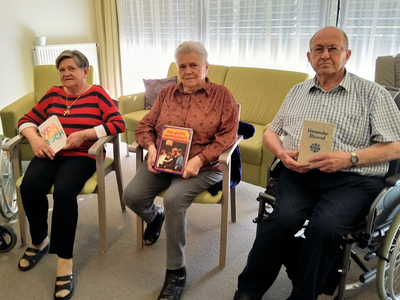 Drei ältere Menschen sitzen in einem hellen Raum auf Stühlen. Jeder hält ein Buch in der Hand. Ein Mann trägt eine Brille und ein kariertes Hemd, während zwei Frauen in farbiger Kleidung neben ihm sitzen. Die Atmosphäre wirkt freundlich und gemeinschaftlich.