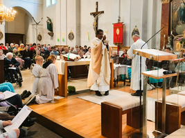 Ein Gottesdienst in einer Kirche mit einer großen Menschenmenge. Ein Priester steht am Altar, während ein weiterer Priester ihm gegenübersteht. Im Vordergrund knien zwei Ministranten, während die Gemeinde aufmerksam verfolgt. Farbige Fenster und religiöse Symbole sind im Hintergrund sichtbar.
