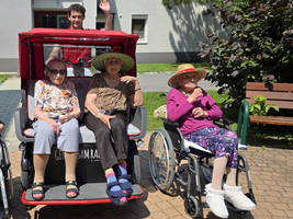 Fünf ältere Frauen in Rollstühlen, vor einer roten Rikscha im Freien.