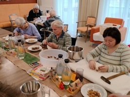Ältere Menschen und eine Frau bereiten gemeinsam Essen in einem Gemeinschaftsraum vor.
